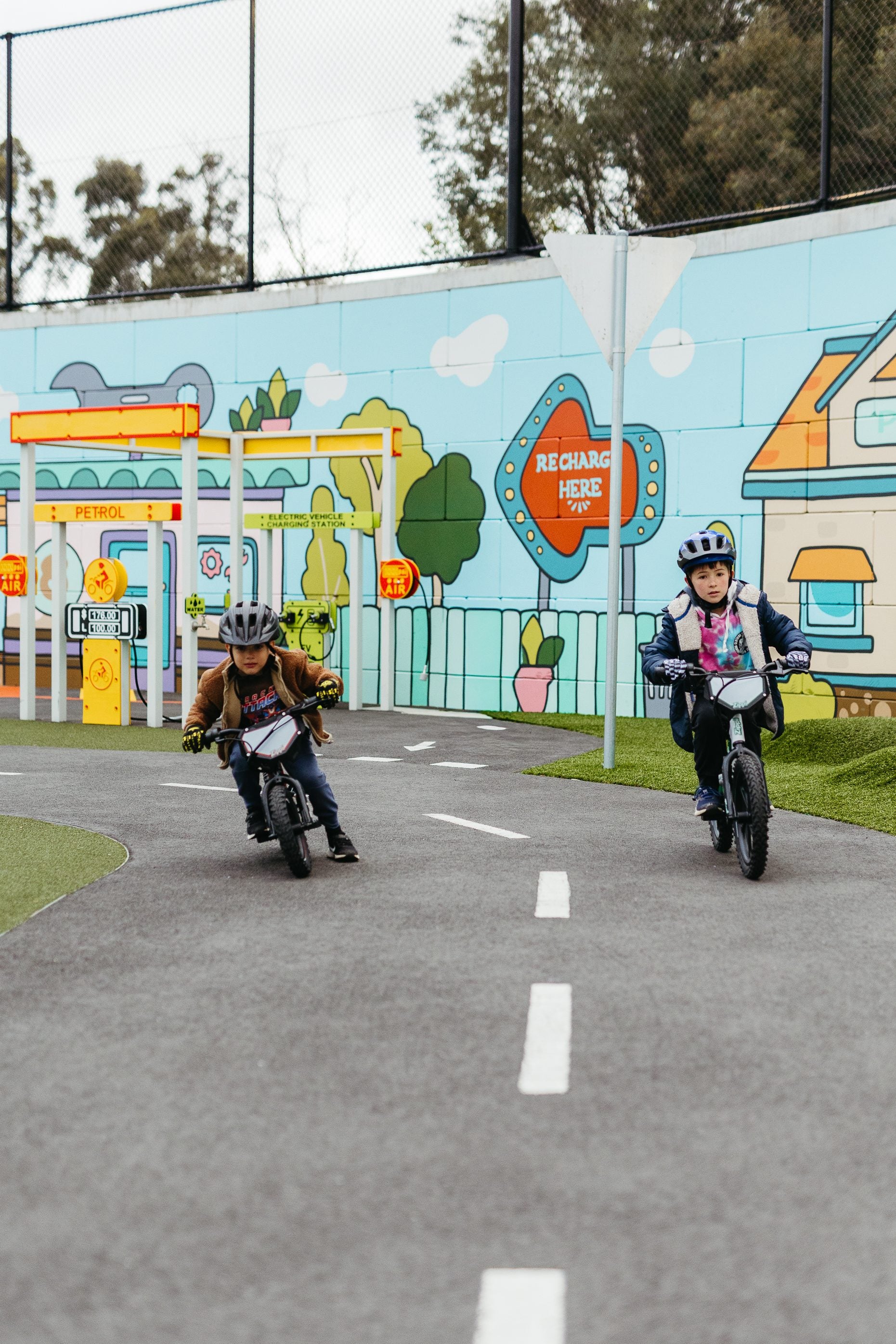 Two children riding Zippi bikes past a colourful 'Recharge Here' mural at a Tasmanian skill park.